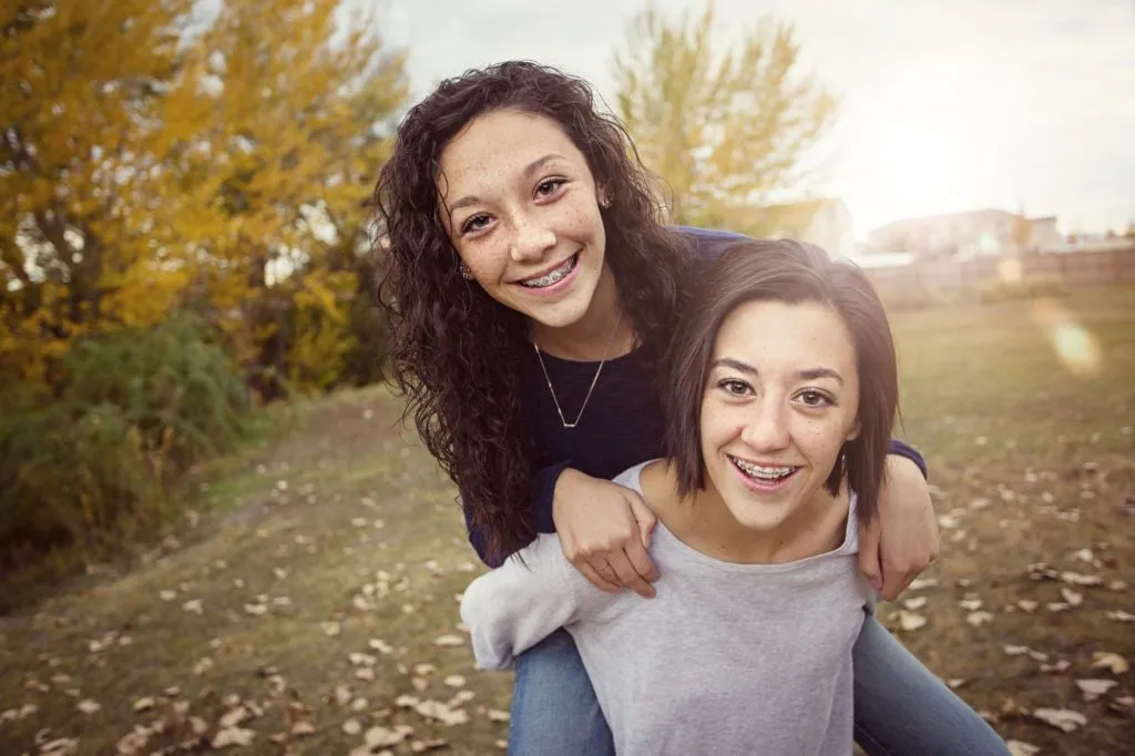 Two smiling teenage girls with braces playing piggyback in a park.