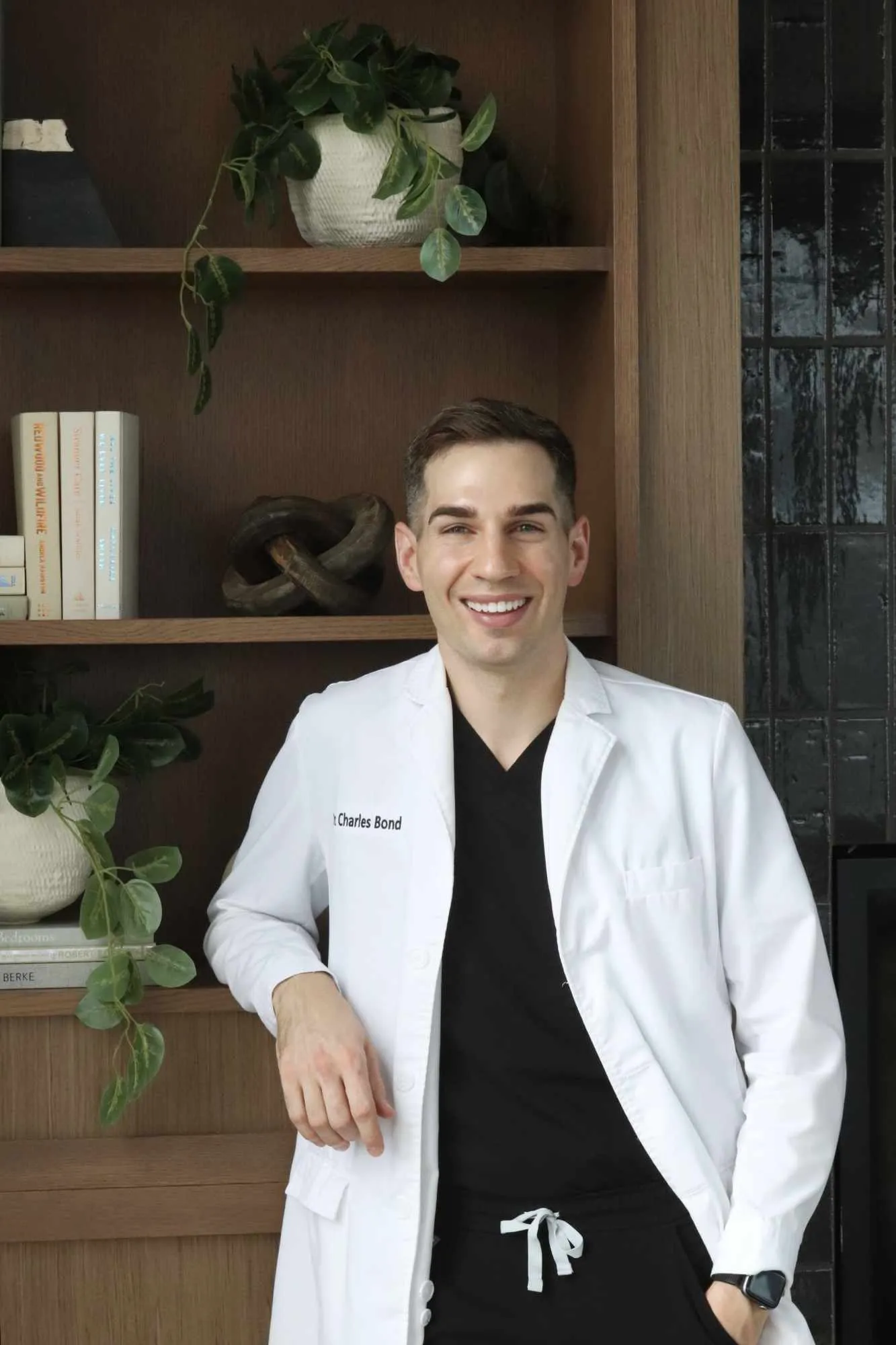 Dr. Charles Bond, orthodontist in Northbrook IL, smiling in a white coat in front of a wood bookshelf at Bond Orthodontics.