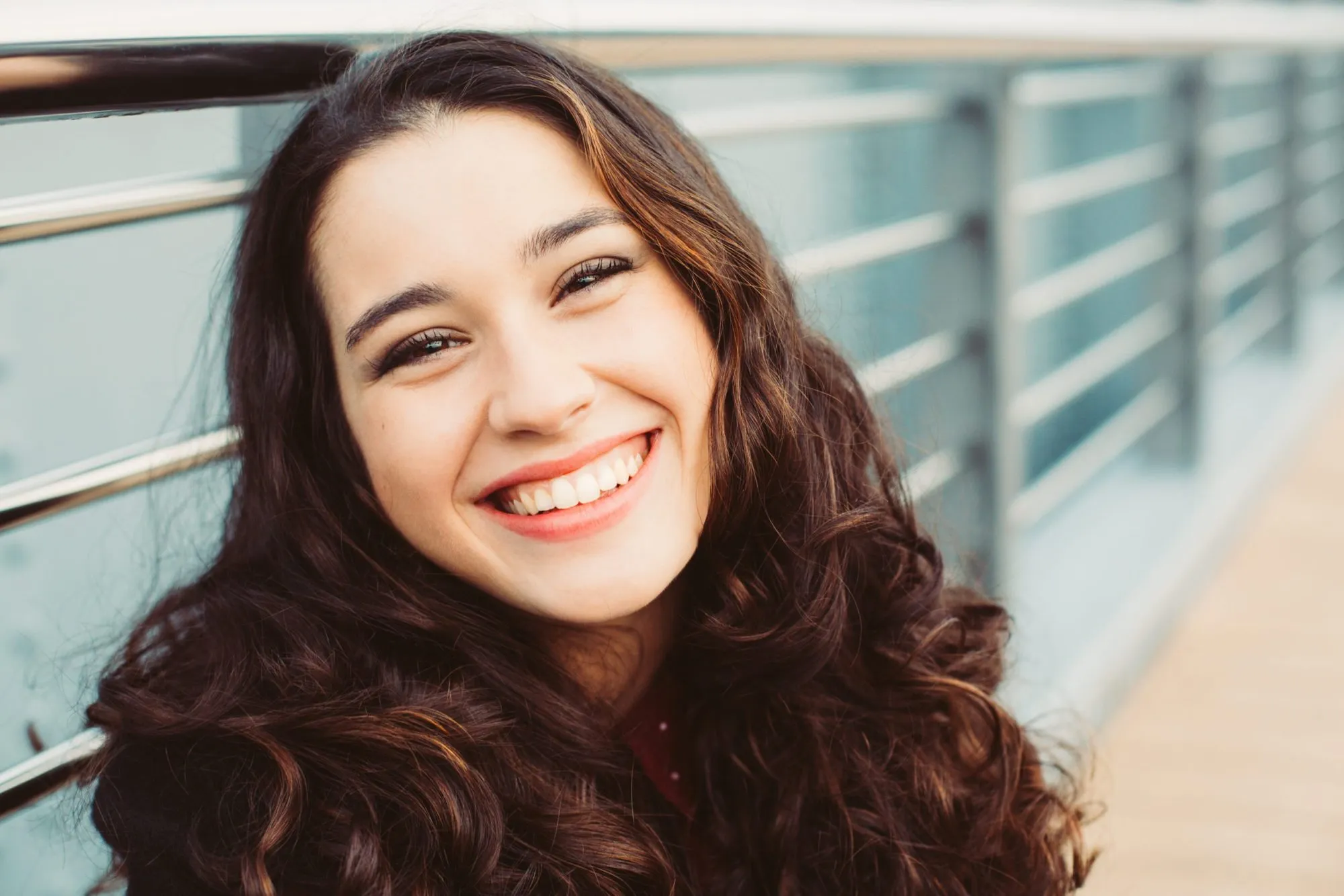 Smiling young woman with long curly hair outdoors by a railing.