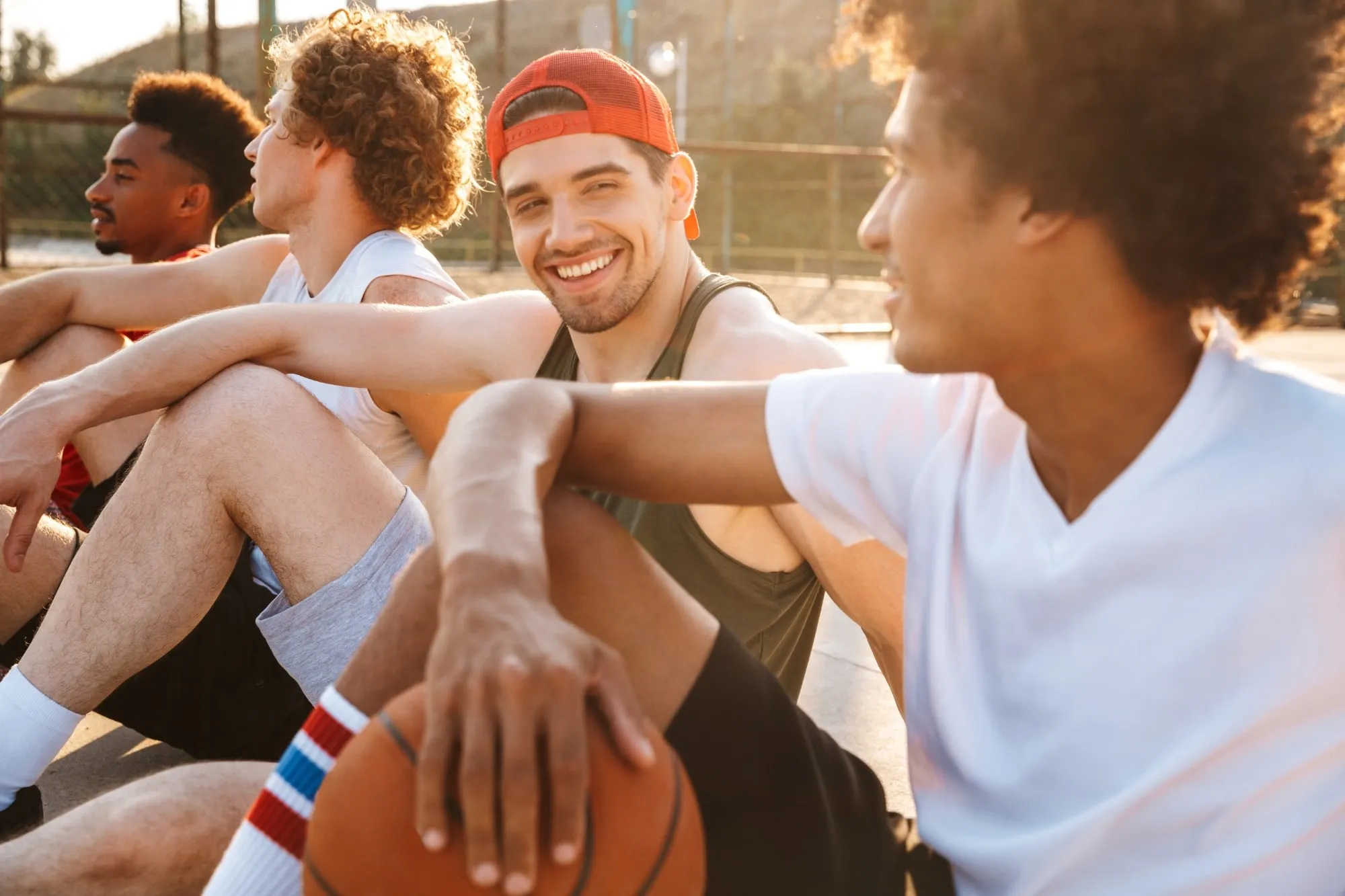 Young men sitting courtside with a basketball on a sunny outdoor court.
