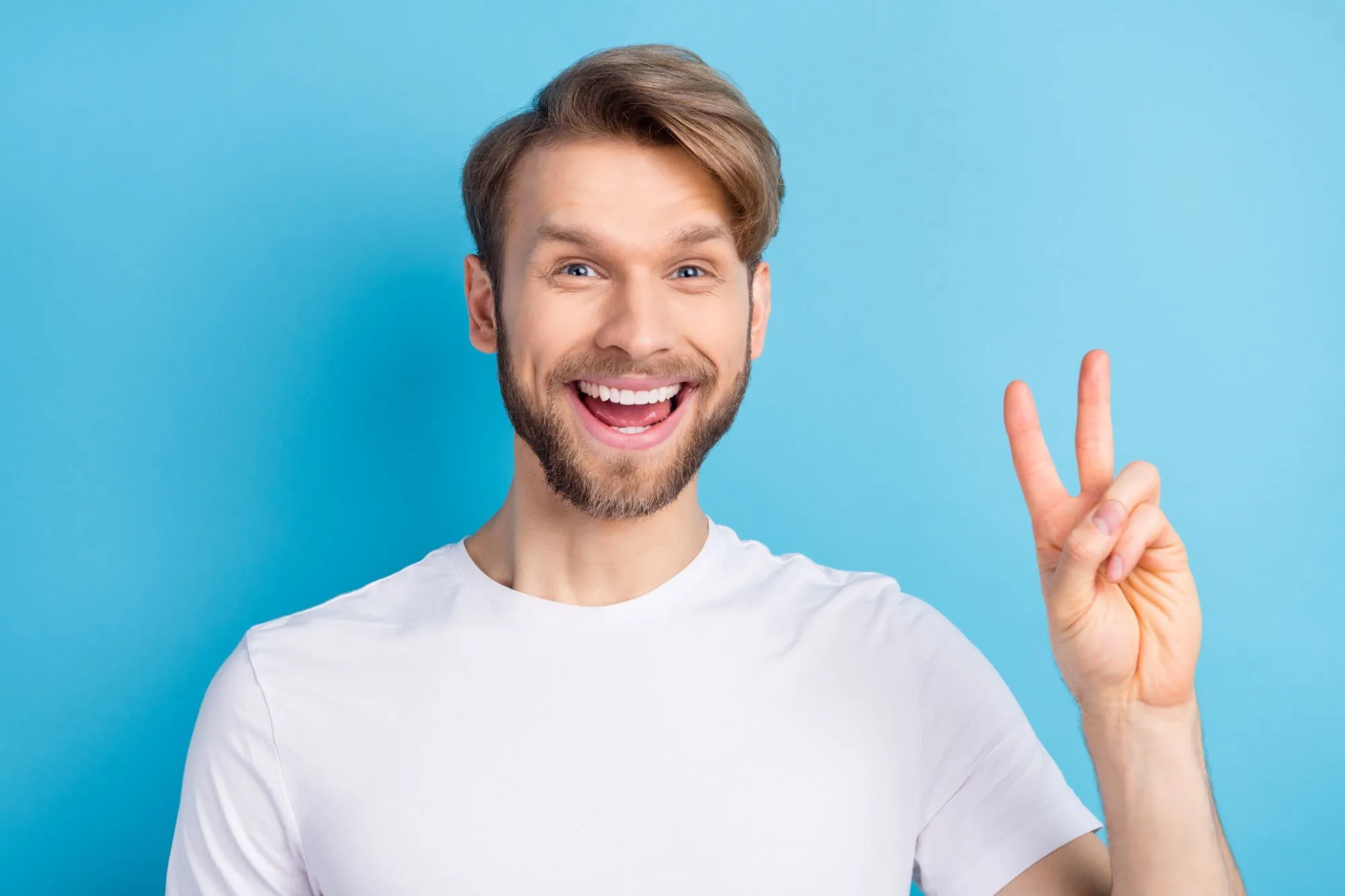 Smiling man in a white T-shirt making a peace sign against a blue background.