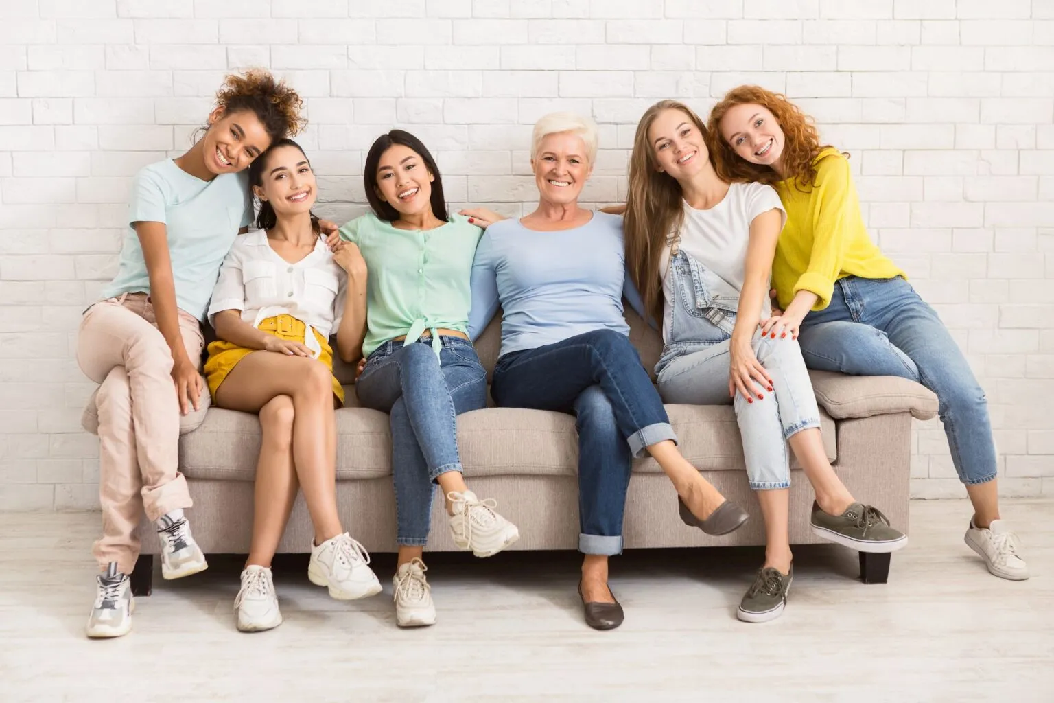 Six smiling women sitting together on a couch against a white brick wall.