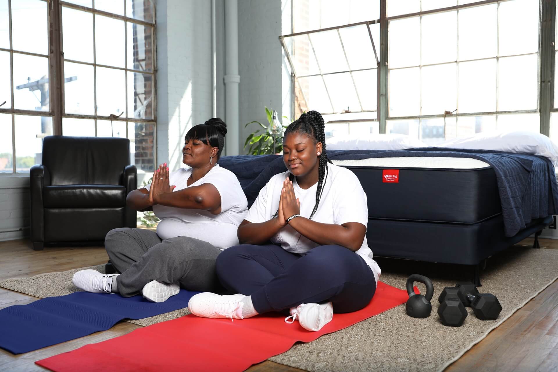 Two women meditating on yoga mats indoors near a bed and dumbbells