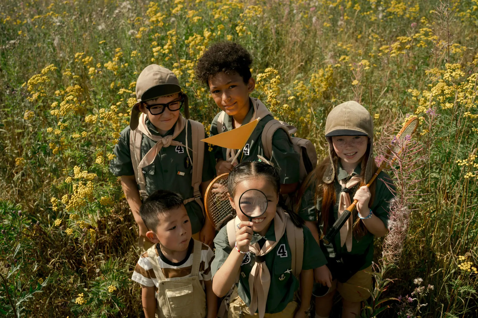 A group of children in scout uniforms exploring a wildflower field, holding tools, and smiling with excitement.