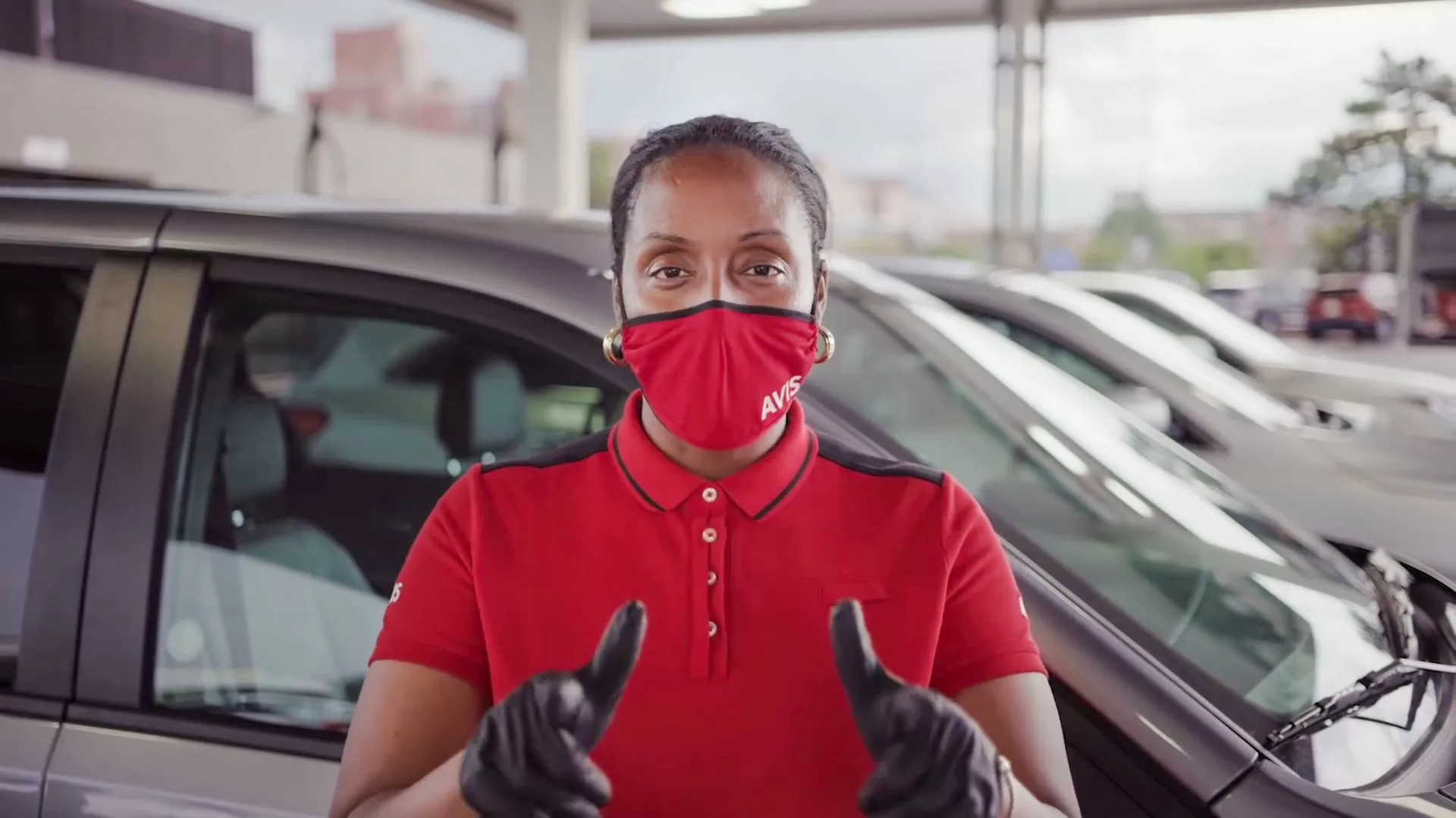 Employee in red uniform and face mask gives thumbs up at car rental parking lot