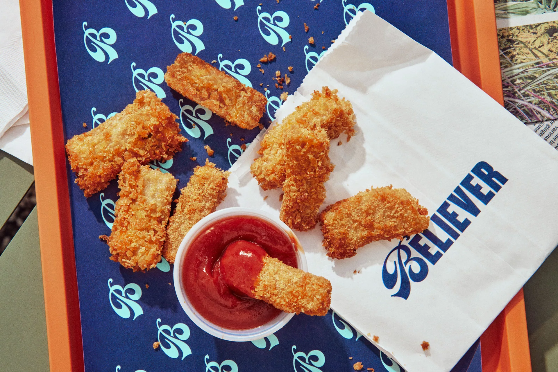 Crispy fish sticks on a blue tray with dipping sauce and a branded paper bag next to them.