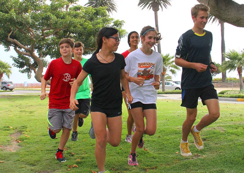 Teenagers jogging together on a sunny day in a park surrounded by trees and palm trees.