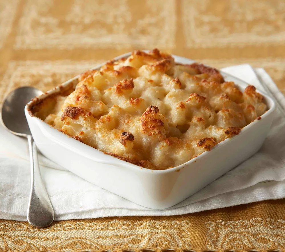 Golden-brown New England lobster mac and cheese in a white baking dish with a spoon on a lace tablecloth.