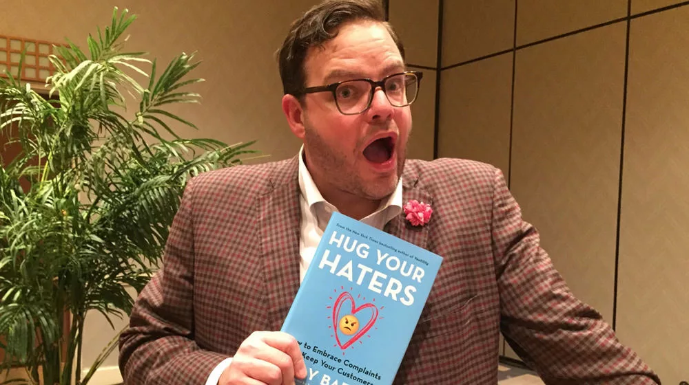 Man in plaid jacket holds "Hug Your Haters" book with an expression of surprise