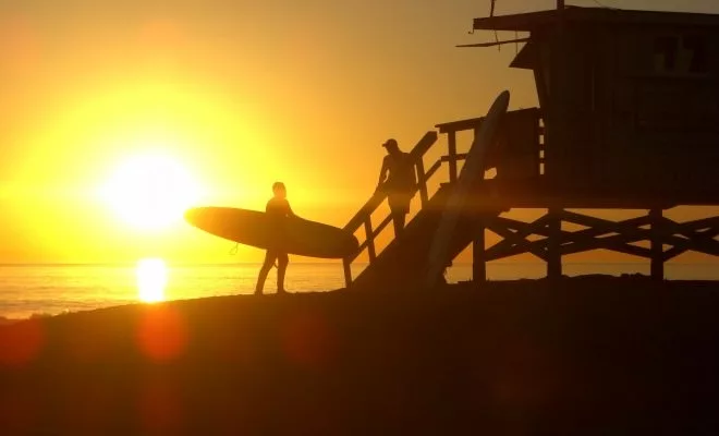 Surfers with boards near lifeguard tower silhouetted against vibrant sunset at beach.