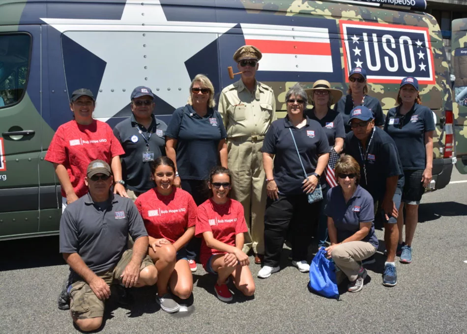 Group of people in USO shirts stand smiling in front of camo-style USO van