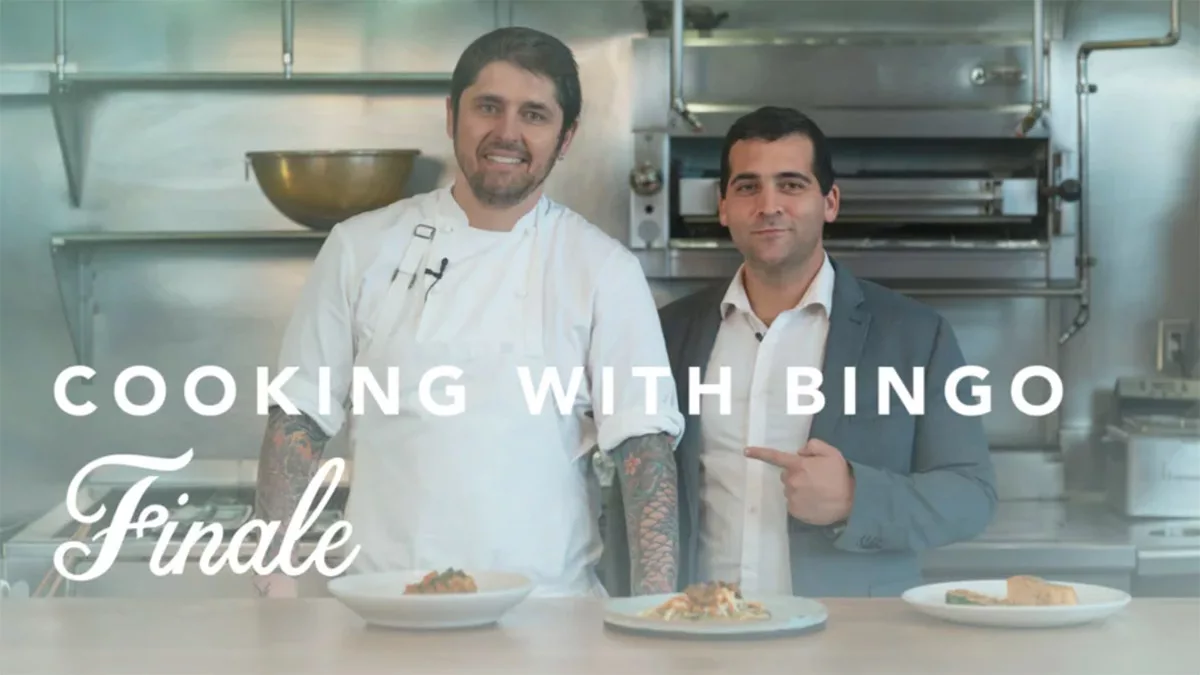 Chef and suited man in a commercial kitchen for "Cooking with Bingo Finale" with plated dishes on the counter.