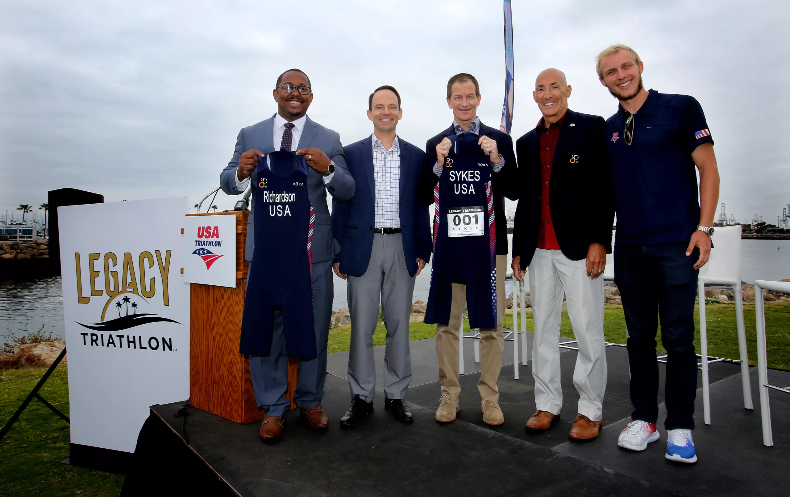 Group of five men holding USA Triathlon suits at Legacy Triathlon event by the waterfront.