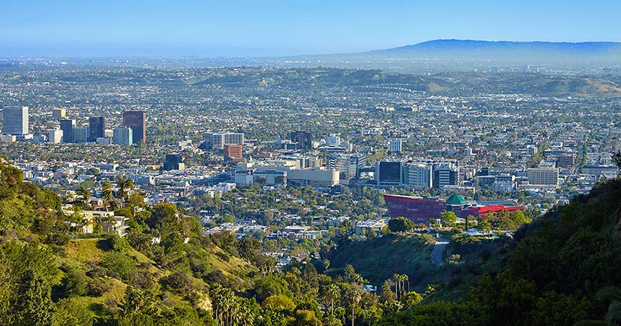 Panoramic view of Los Angeles skyline with lush greenery in the foreground and sprawling cityscape under a clear blue sky.
