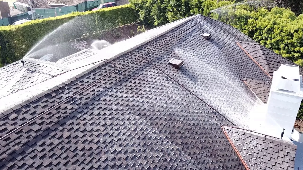 Rooftop with dark shingles being cleaned with water spray under bright sunlight and surrounded by green trees.