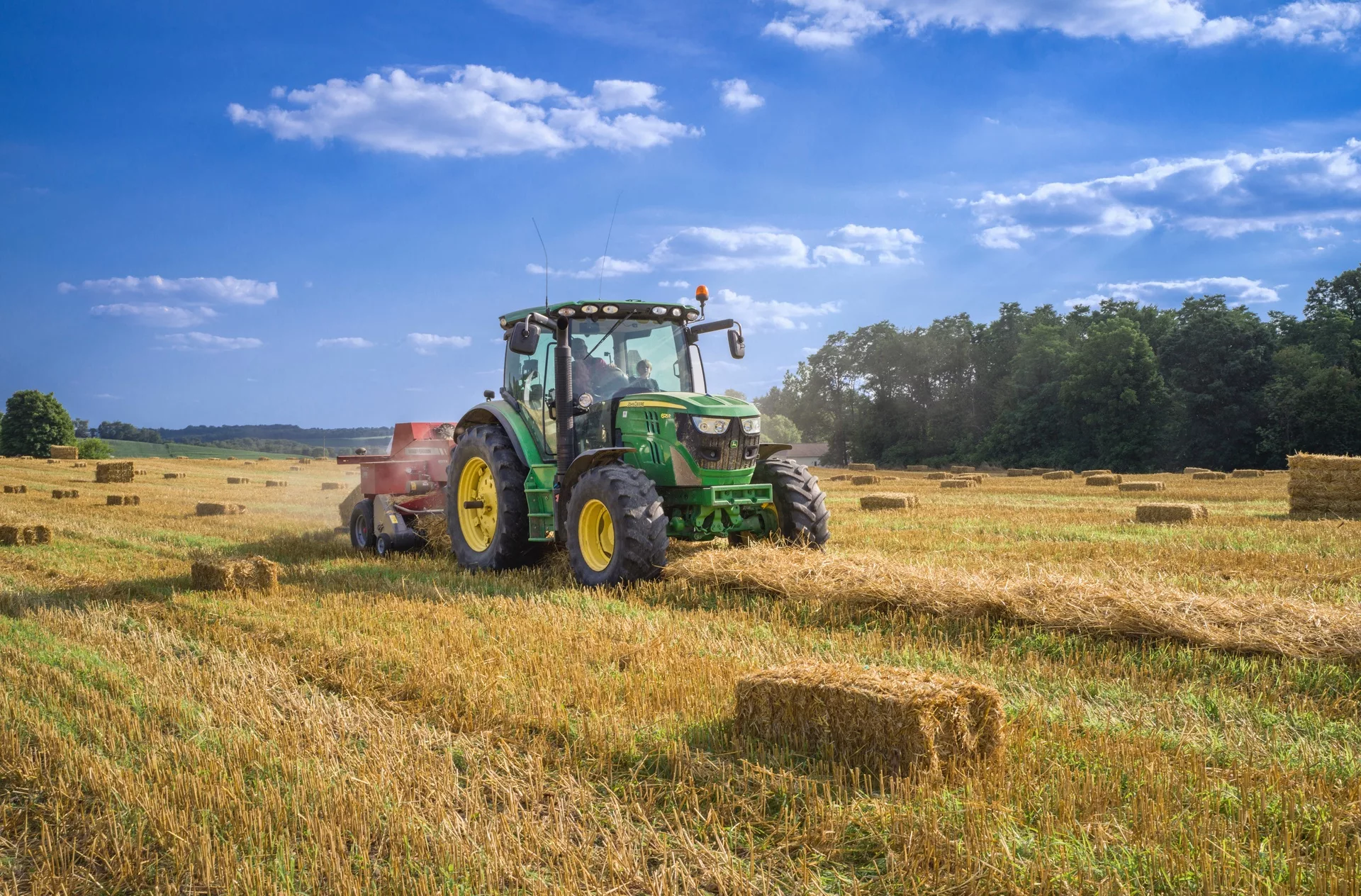 Green tractor baling hay in sunny field; blue sky and scattered clouds in background.