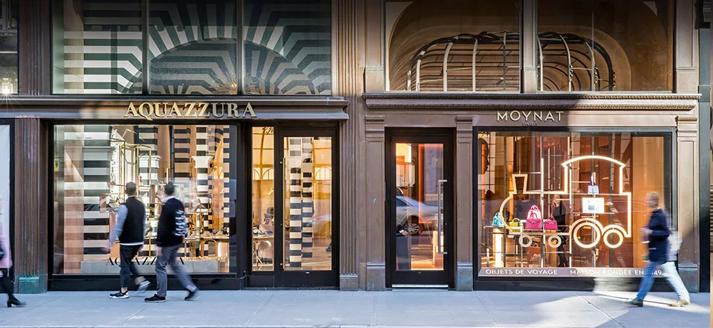 Two fashion store fronts with geometric window displays and pedestrians walking past on a city street.