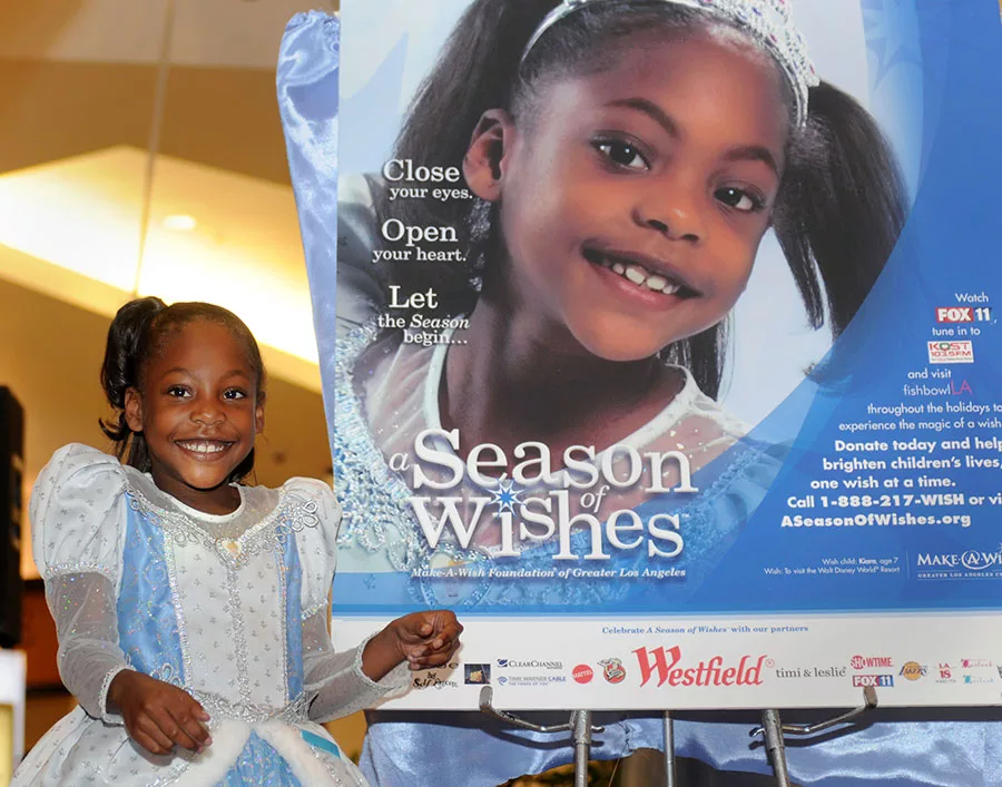 Smiling girl in a blue dress poses beside "A Season of Wishes" poster