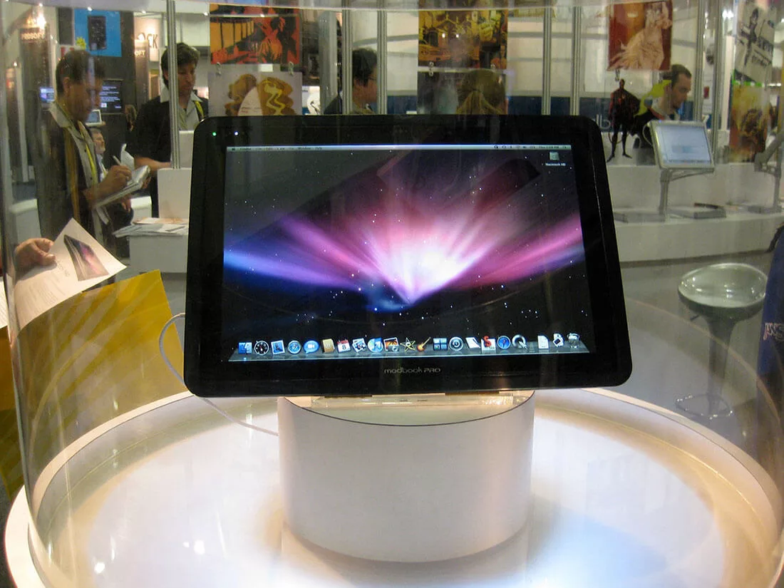 Tablet prototype displayed on a round stand with attendees in the background at a tech conference.