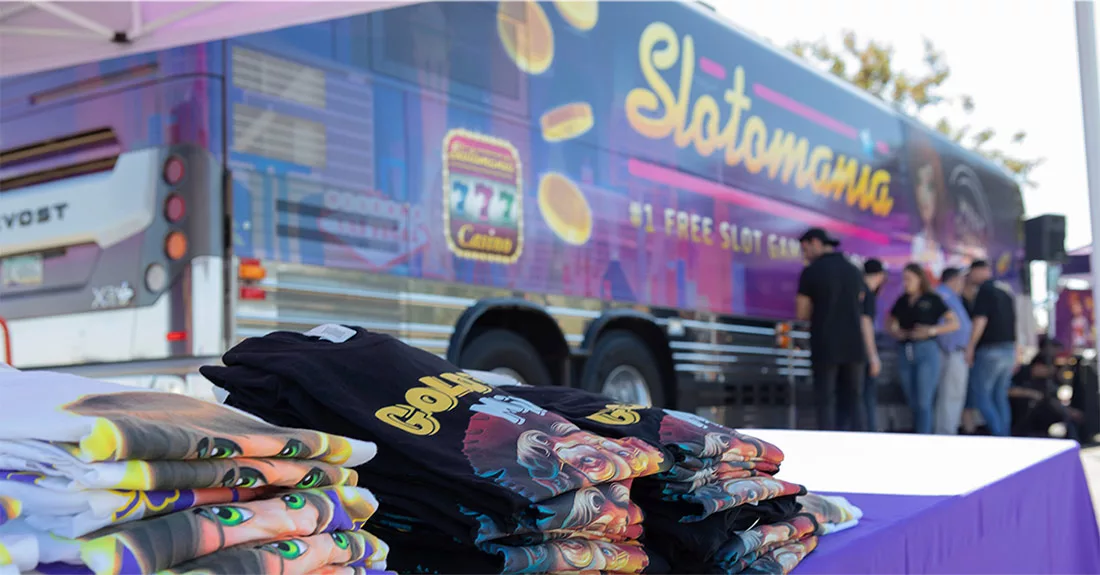 Stacked Slotomania shirts with vibrant bus backdrop at outdoor event.