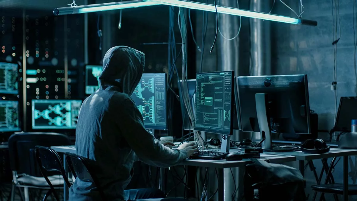 Hacker in a dark room working on multiple computer screens with code visible