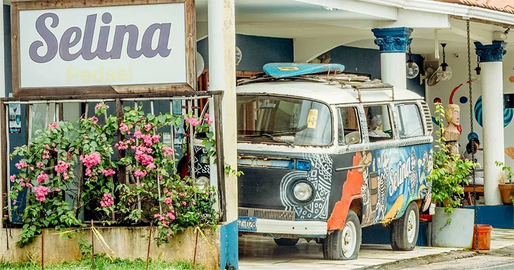 Colorful vintage van with surfboard parked beside pink flowers and a "Selina Pedasi" sign outside a building.