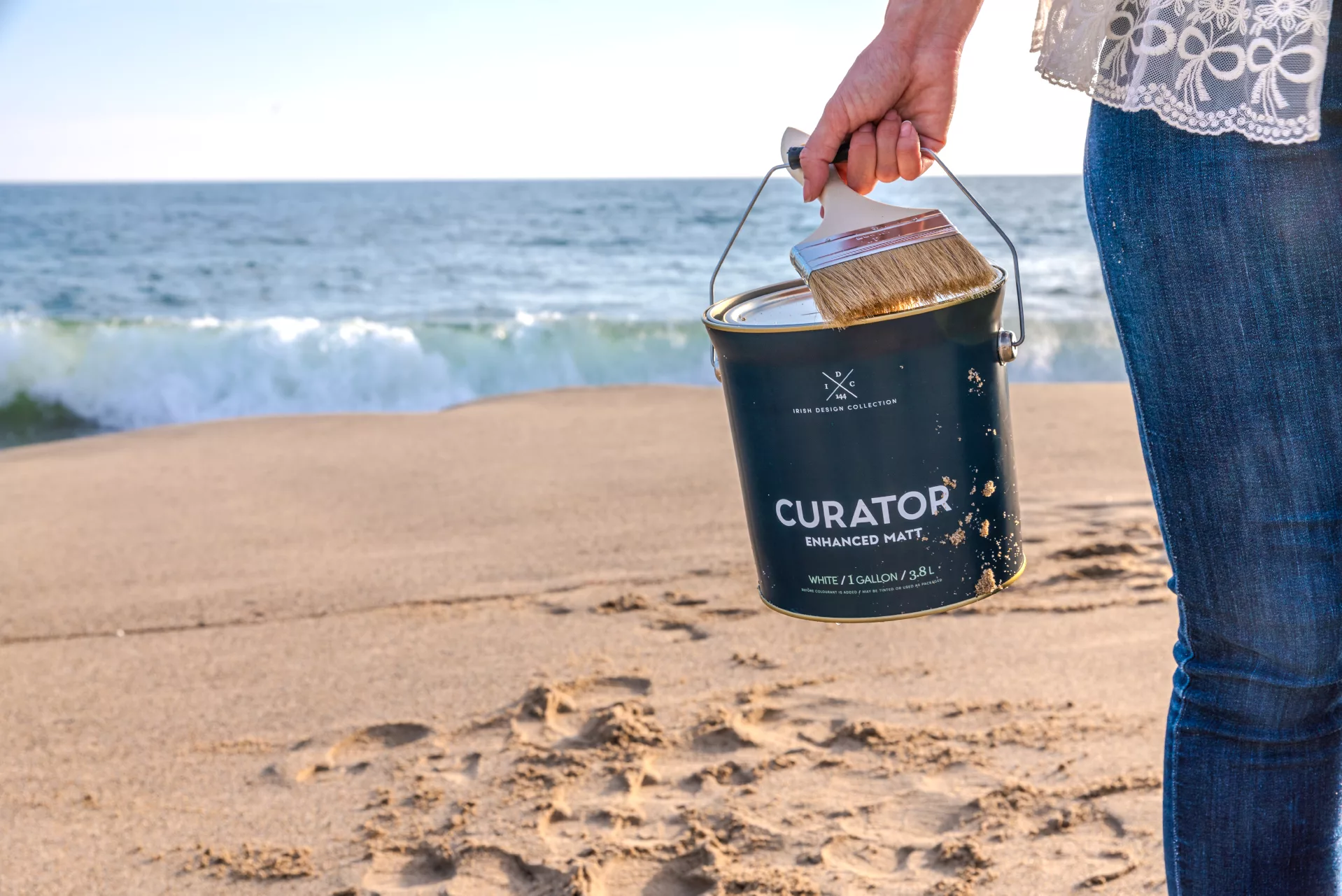Person holding a paint bucket and brush on a sandy beach with ocean waves in the background.