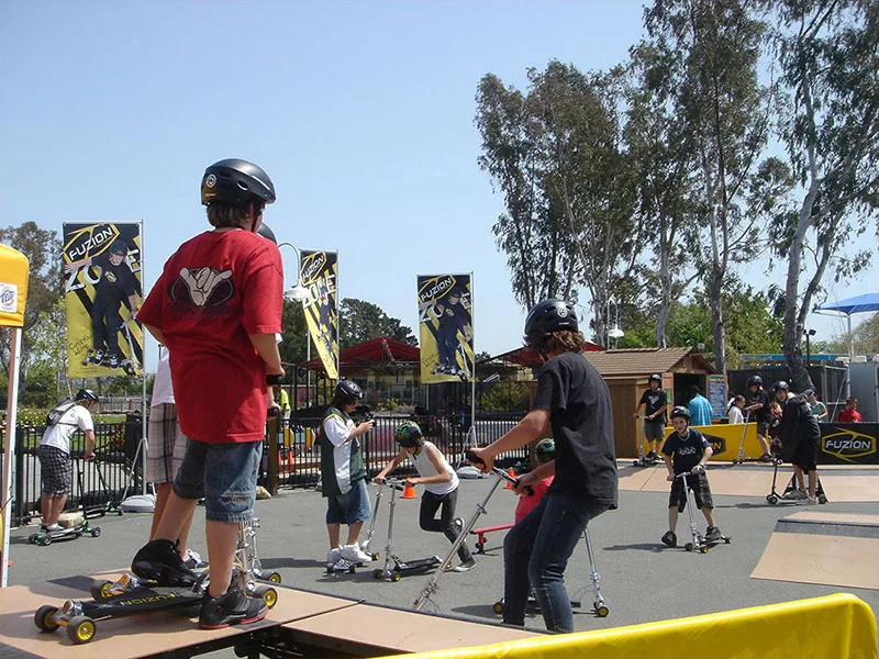 Kids in helmets ride scooters at an outdoor skate park
