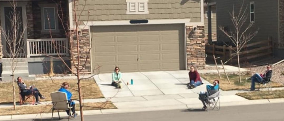 Neighbors enjoying a sunny day outdoors, sitting in lawn chairs six feet apart, in front of suburban homes.