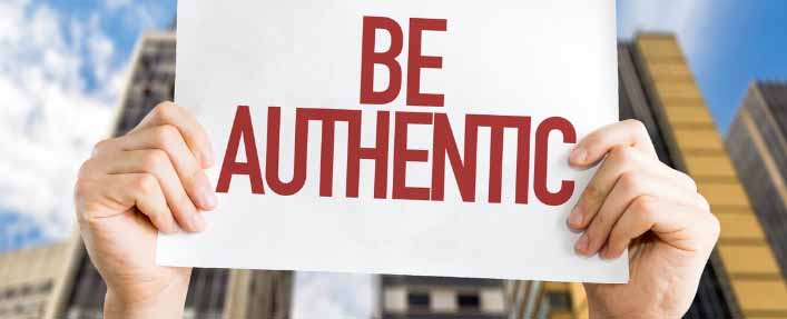 Hands holding a sign with "Be Authentic" against a cityscape