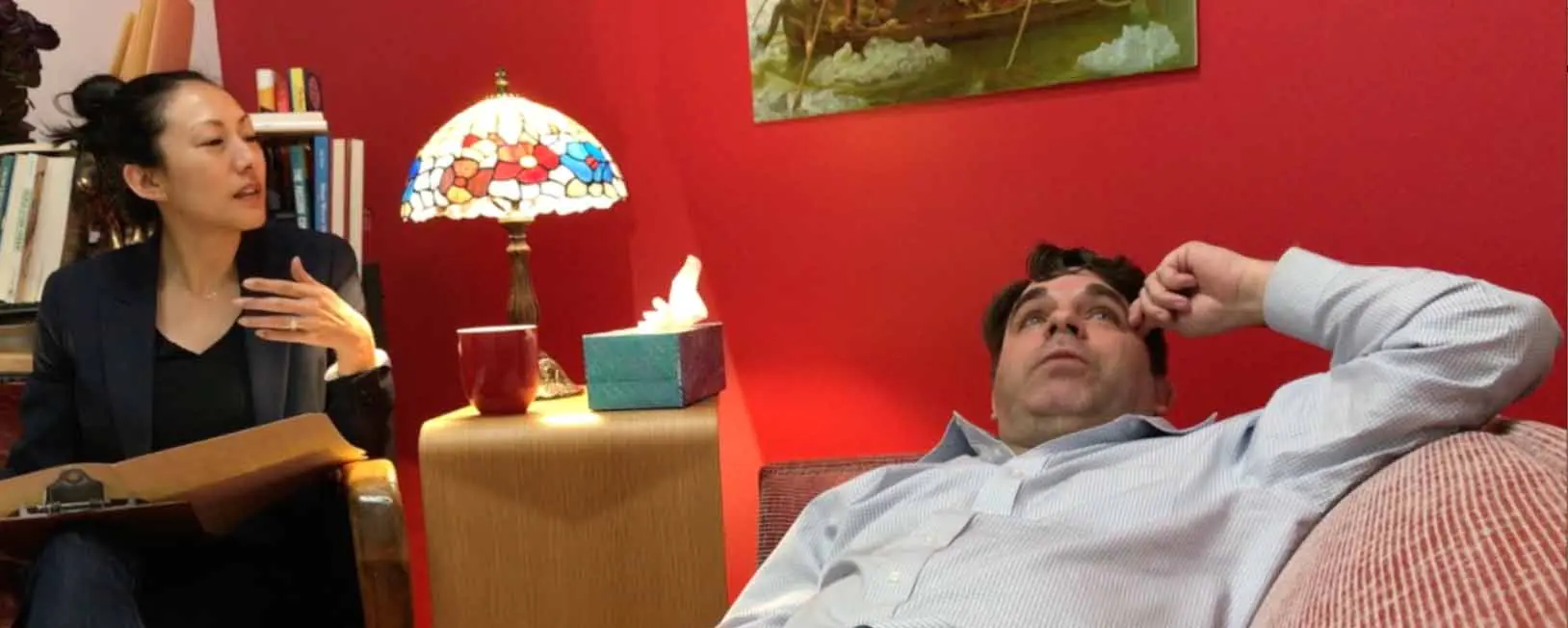 Therapist with clipboard converses with man on couch in room with red walls
