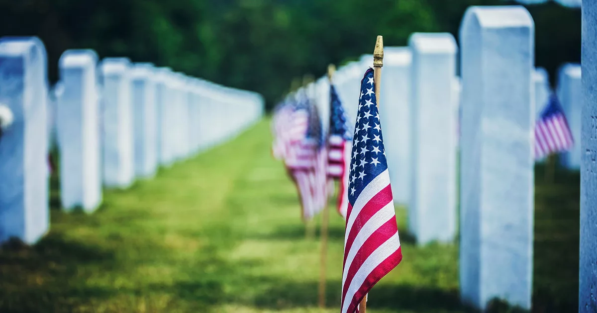 American flags lined up in a cemetery with white headstones on a sunny day