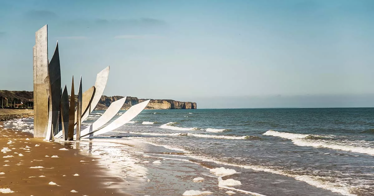 Metallic sculpture on Omaha Beach with ocean waves and distant cliffs under blue sky.