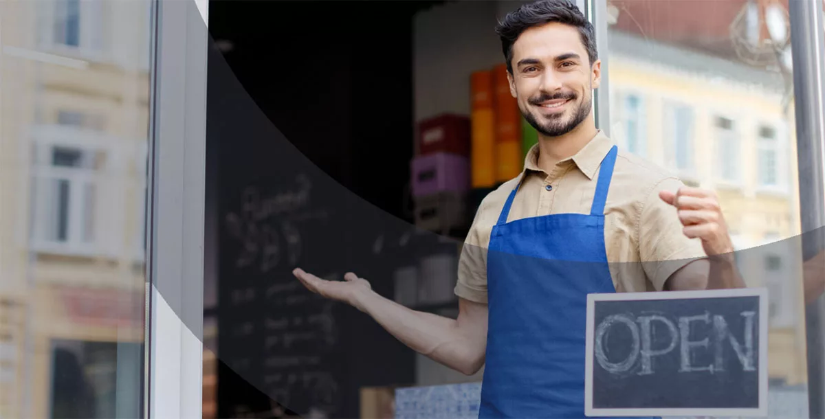 Smiling male shop owner in blue apron welcomes customers with open sign at storefront.