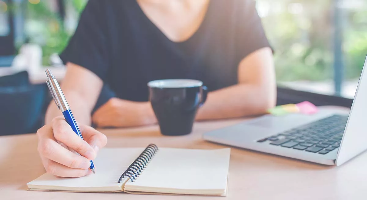 Person writing in a notebook with pen beside a laptop and coffee mug on a table.