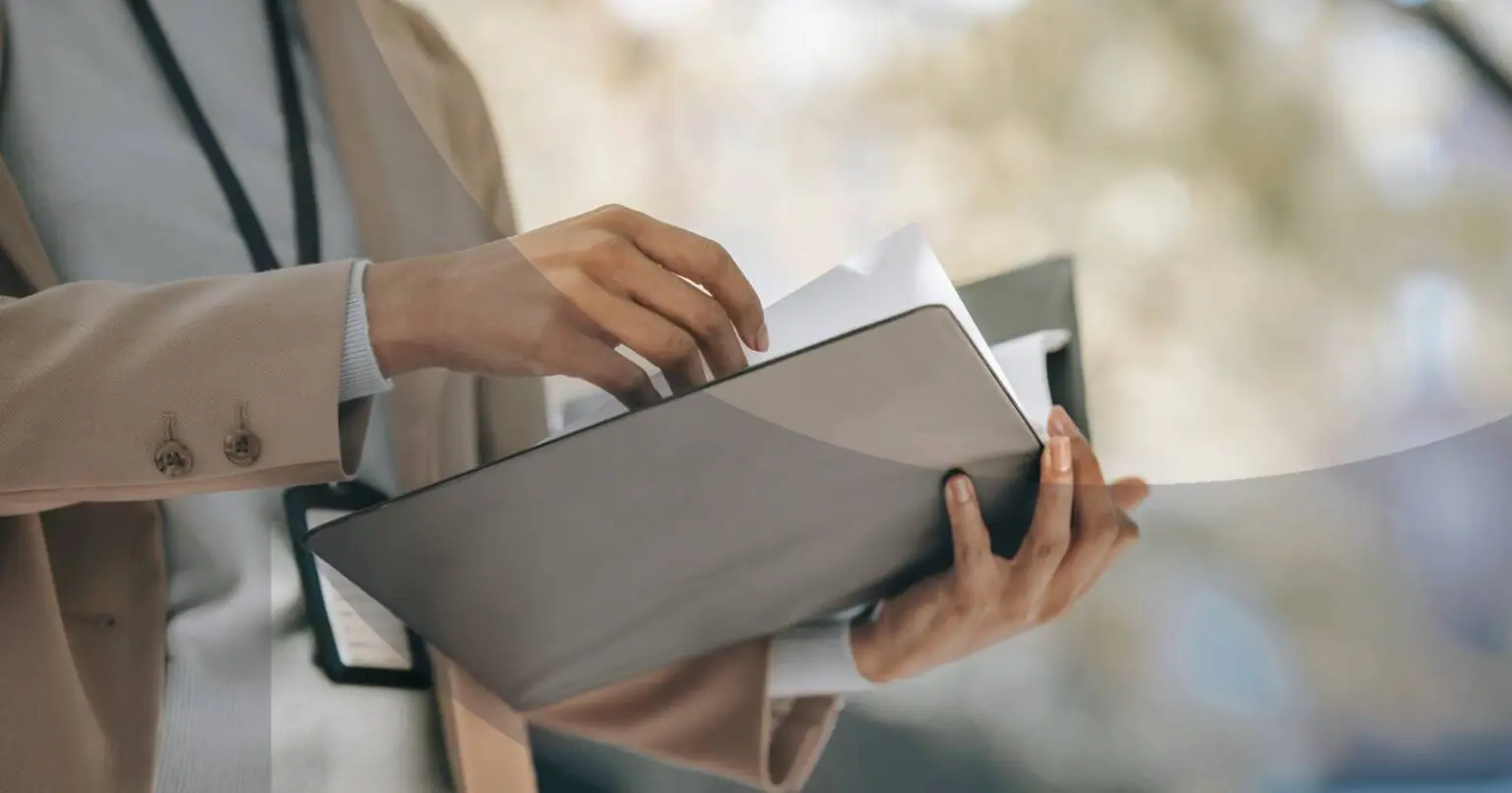 Person in beige blazer holding and sorting documents in a folder outdoors.
