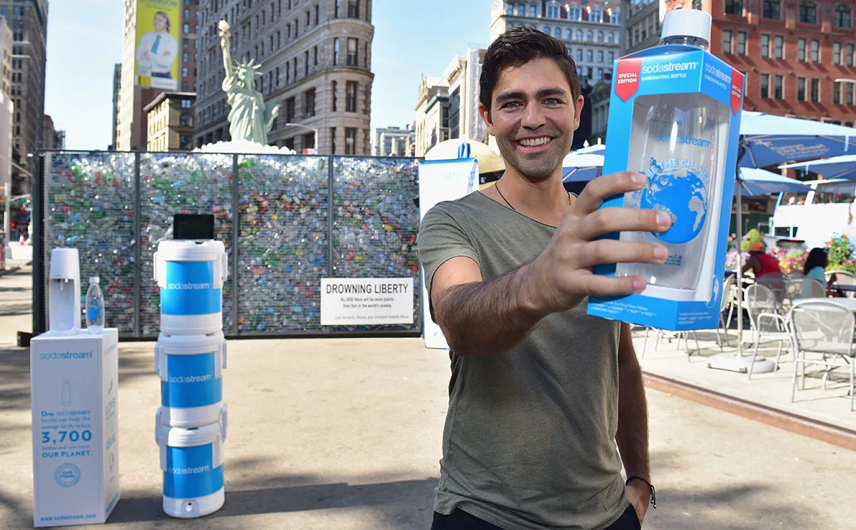 Man holding SodaStream bottle in front of plastic-filled Liberty statue backdrop at outdoor event