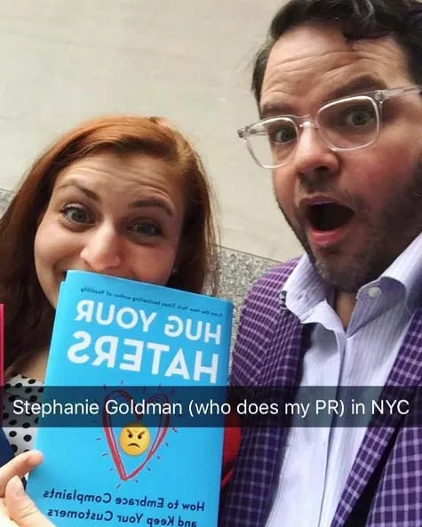 Two people smiling with a "Hug Your Haters" book
