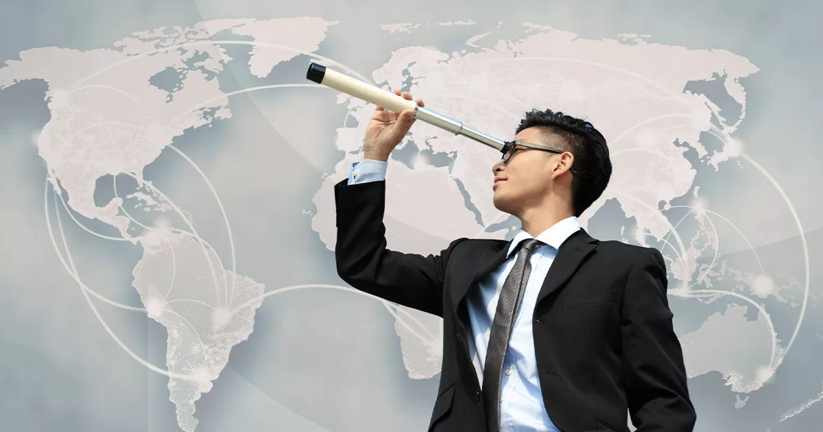 A businessman in a suit looks through a telescope against a digital world map background