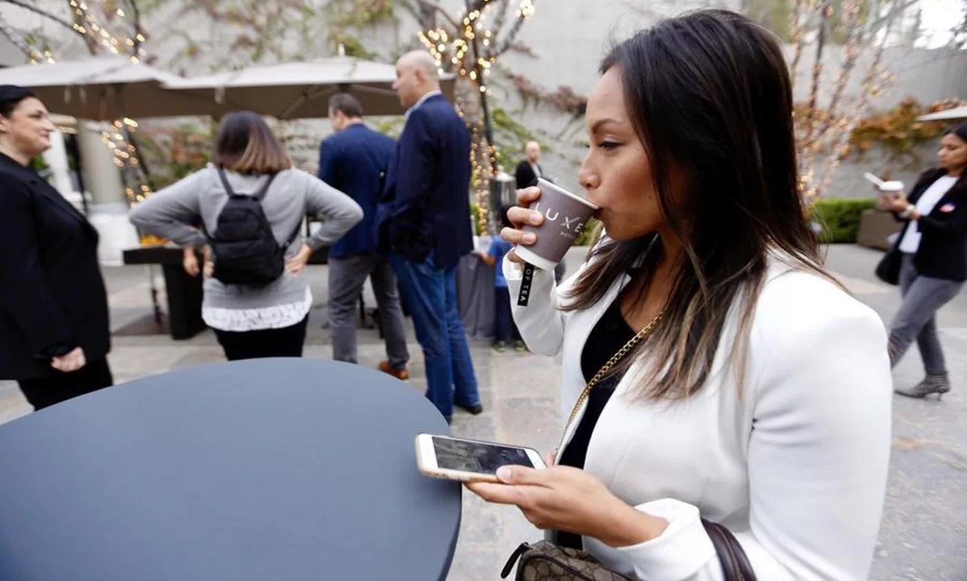 Woman in white blazer sipping coffee and holding a smartphone at an outdoor networking event.