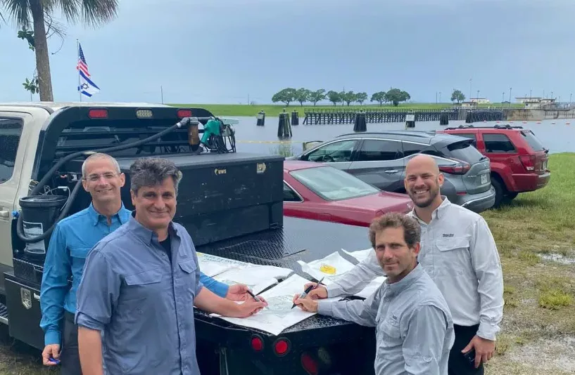 Group of four engineers working on blueprints at a truck's tailgate near a river