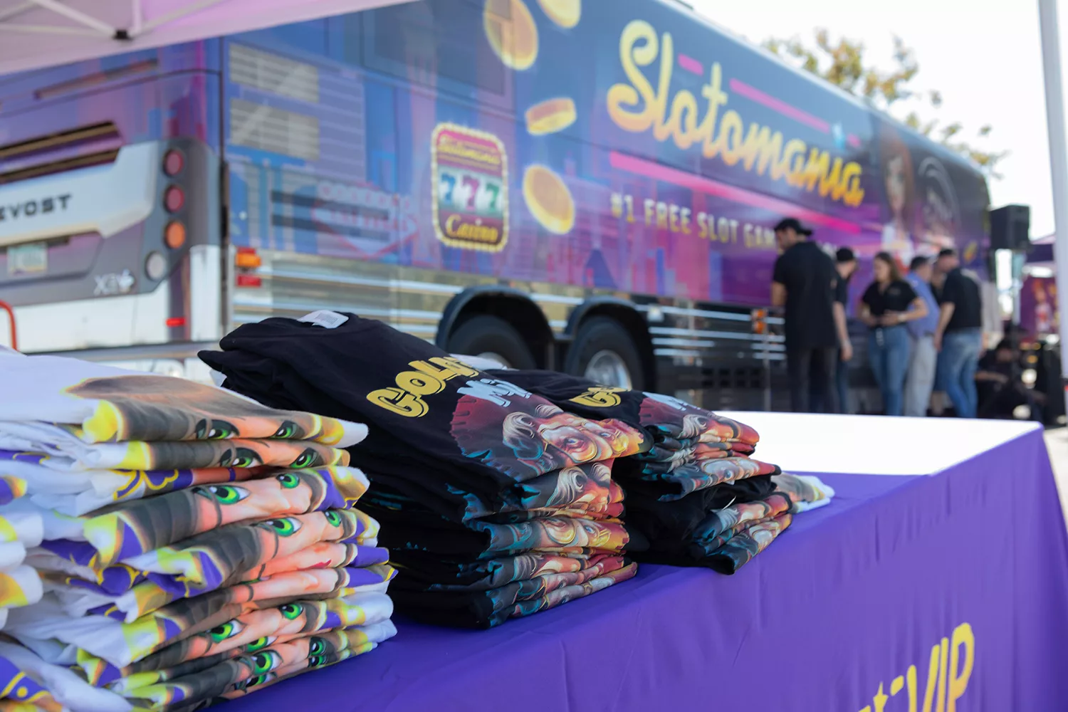 Stacks of colorful Slotomania-themed shirts on a table in front of a branded Slotomania bus at an event.