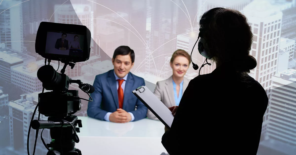 TV studio setup with two news anchors on camera and a director reviewing notes.