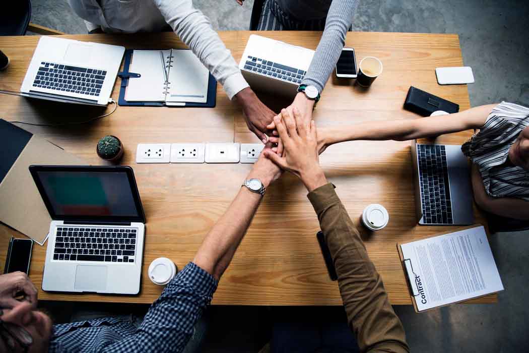 Teamwork concept with five people joining hands over a table with laptops and papers in an office setting.