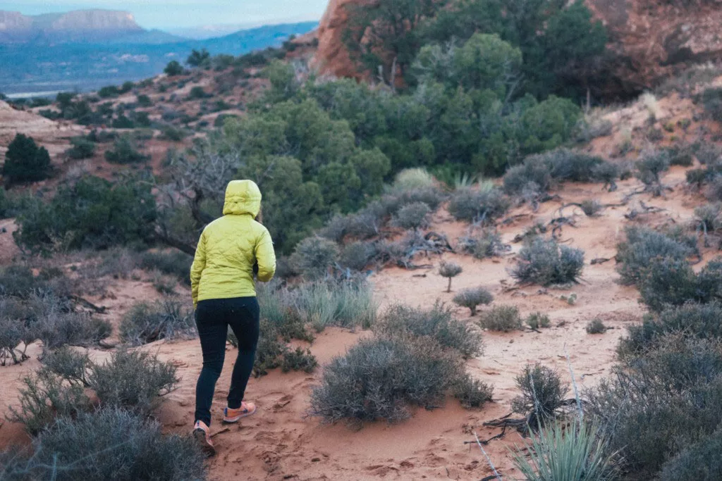 Person in a yellow jacket hiking through a rocky desert trail surrounded by green shrubs and distant mountains.
