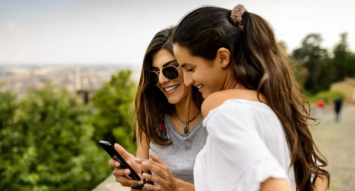 Two women smiling and looking at a smartphone outdoors with nature in the background.