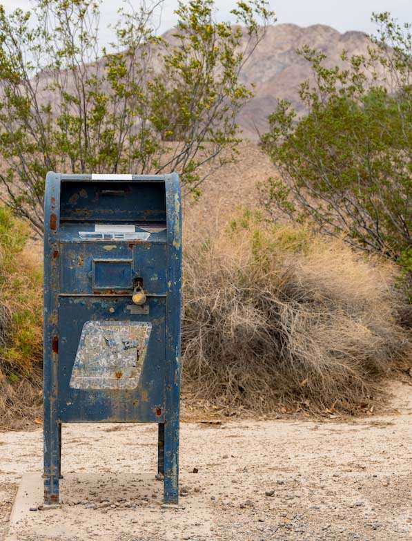 Rusty blue mailbox in a desert landscape with mountains and scrubby bushes in the background.