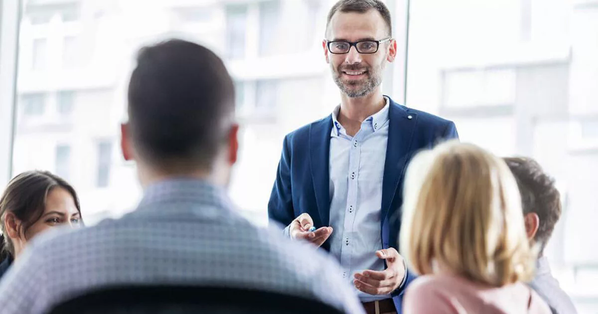 Business meeting with a smiling man in a blue blazer presenting to colleagues in a bright conference room.