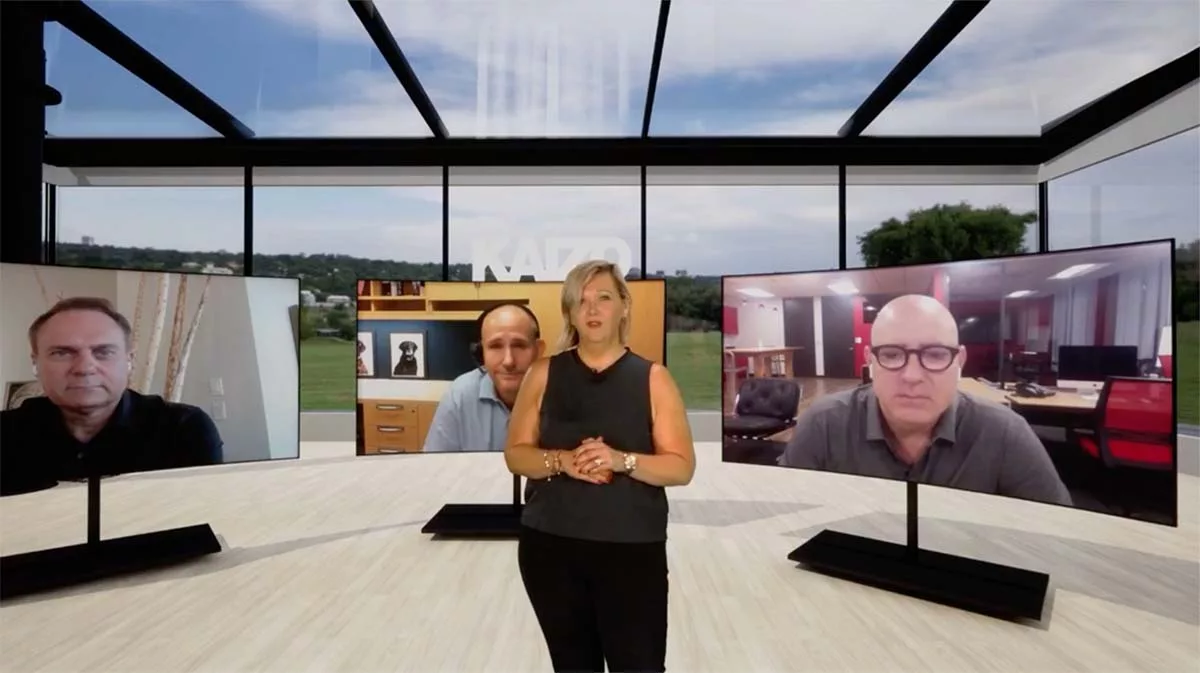 Woman hosting a virtual meeting with three men on screens in a modern office with large windows.