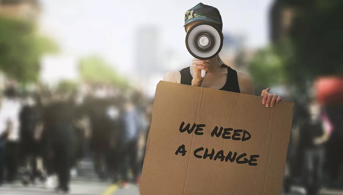 Protester holding a megaphone and cardboard sign reading "We Need a Change" in a crowd during a peaceful demonstration.